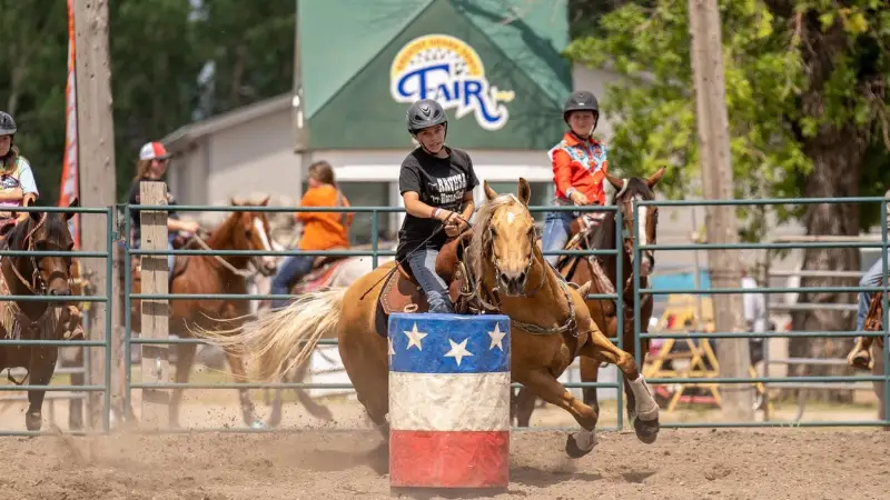 Grand-Forks-4H-Club Grand Forks County Fair hosts barrel racing around American flag stripped barrel. Girl ridding a horse in the arena.