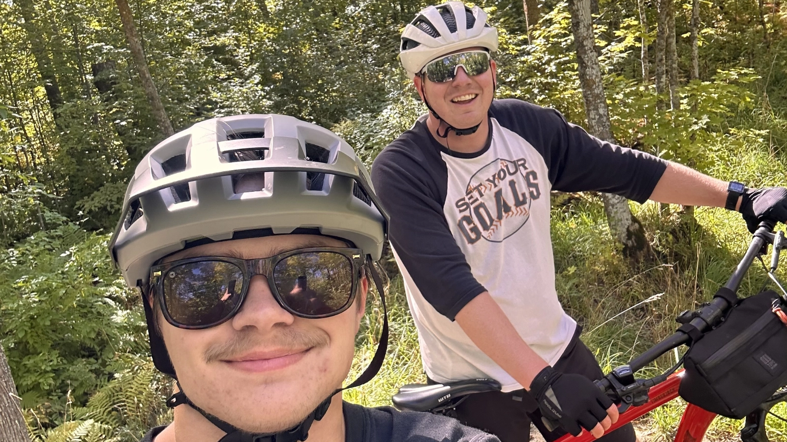 selfie-biking WCT writer Paul and a friend wear sunglasses and a helmet as they smile and bike through a heavily wooded area.