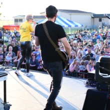 Grand Forks County Activities A guitarist and a singer face away from the camera as they performing for a crowd of hundreds.
