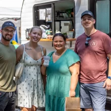 Community for all Picture of four adults at the downtown market in the summertime. Two men on the outsides and two women on the insides. Smiling at the camera in front of a little camper.