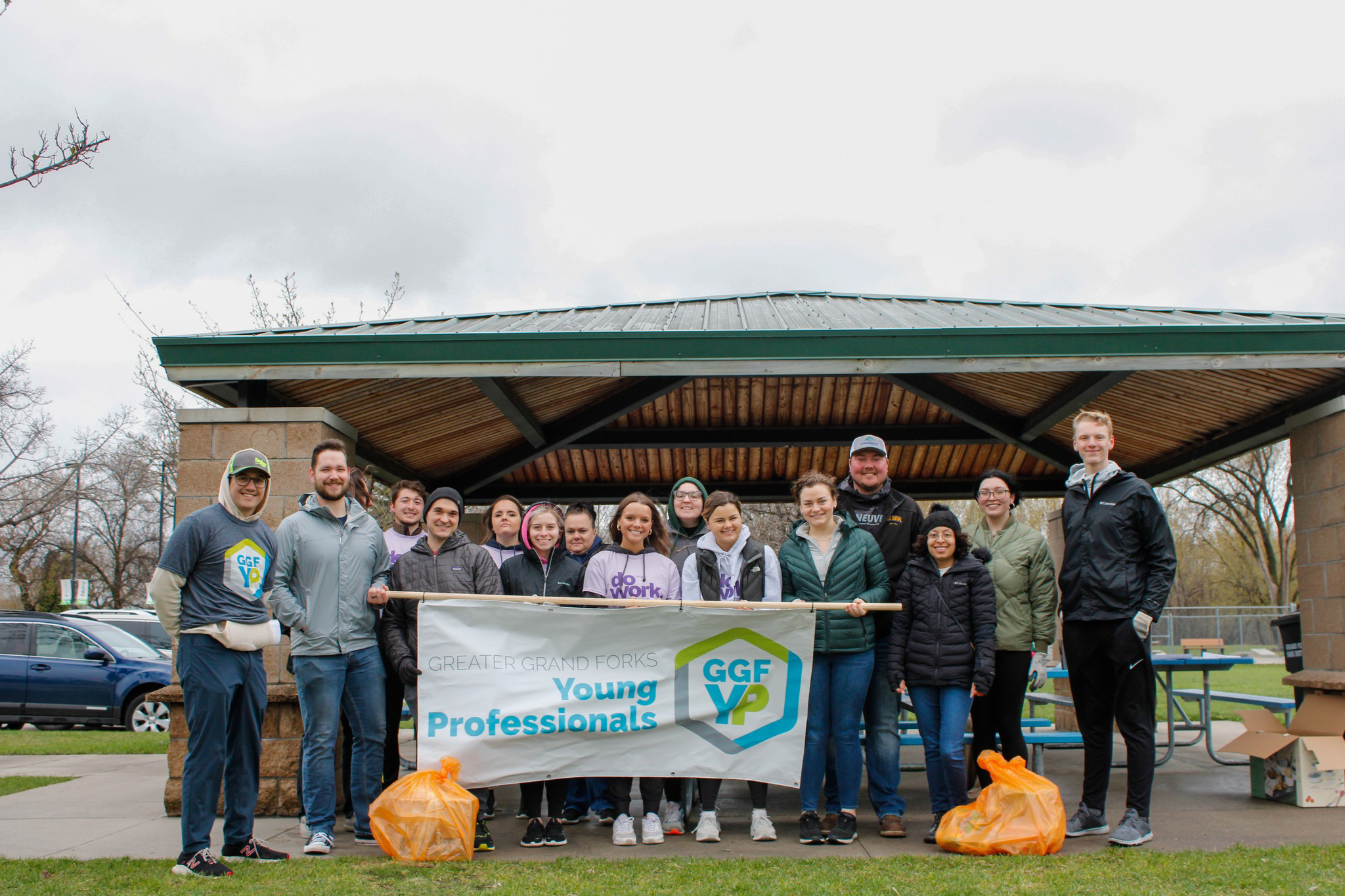 Orange Bag Volunteering Group Photo Young Professionals pose in a group with the GGFYP Banner for the Orange Bag Donations event. They are posing in front of an outdoor shelter in a park in Grand Forks on a dreary day.
