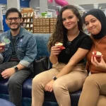 Author Adrian sits at the GF Public Library alongside two women holding red velvet cupcakes and smiling.