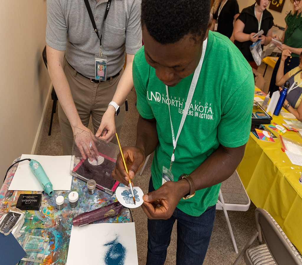 Art Volunteering InternGF A UND student is pictured participating in an art volunteering event through the InternGF Summer Cohort series. He wears a bright green UND shirt and his holding his art piece delicately while painting it.
