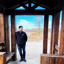 Ben article one featured image Author Ben stands under a wooden archway with a clear blue sky behind him as he smiles off to this distance.