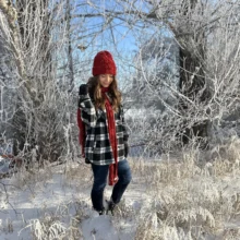 Natalie Winter Featured Image Author Natalie wears a black and white flannel with a matching red hat and scarf, as she stands in a scenic frosty winder landscape.