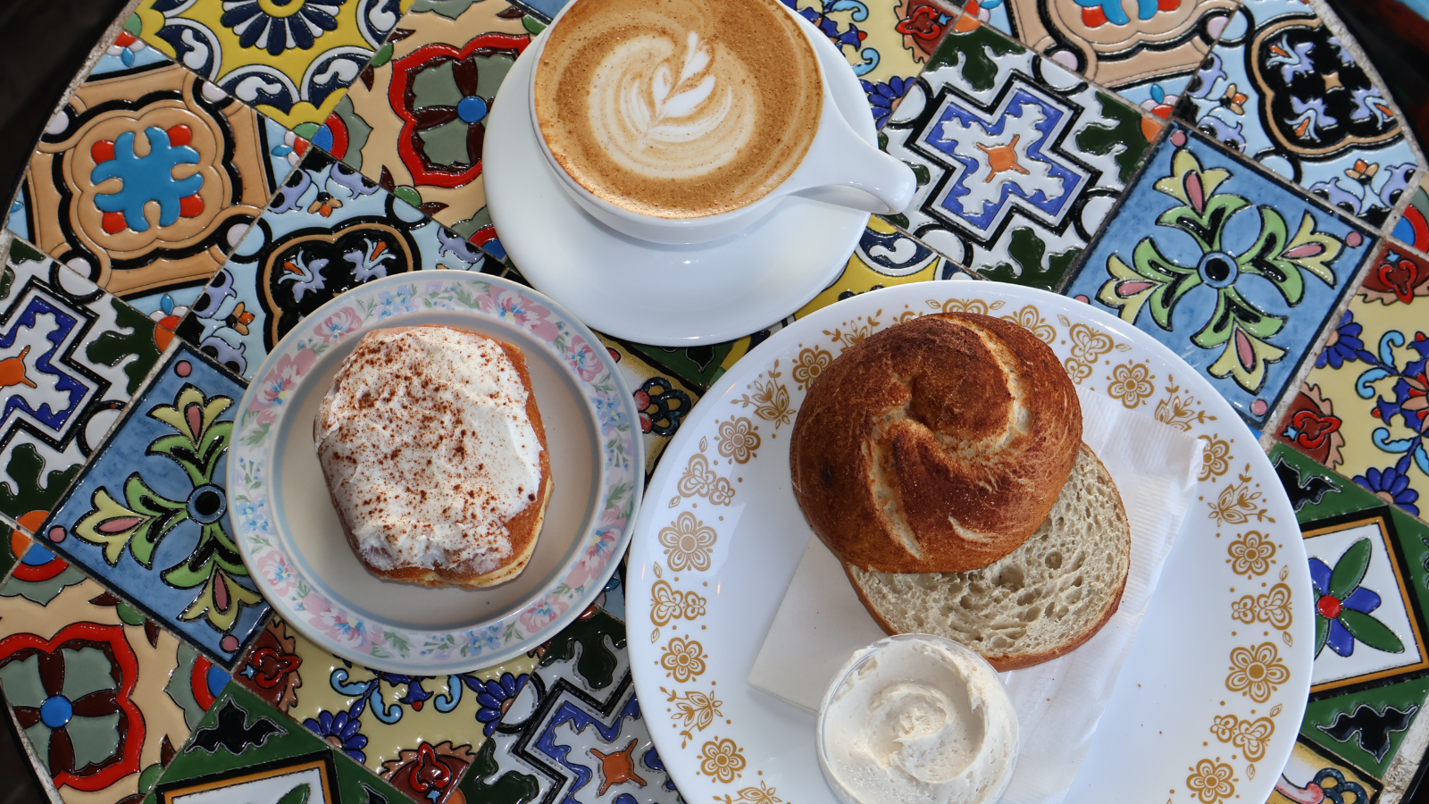 Urban Stampede Food Image An overhead image from Urban Stampede. Showcasing a latte, bagel, and cinnamon roll on a mosaic tiled table. yum!