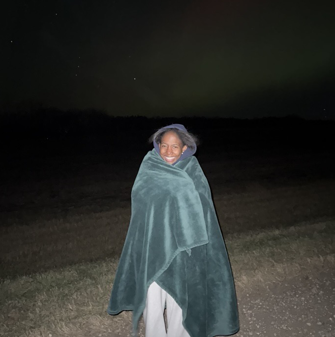 Author Precious stands and smiles at the camera during a northern lights chasing trip while she's wrapped up in a cozy blanket!